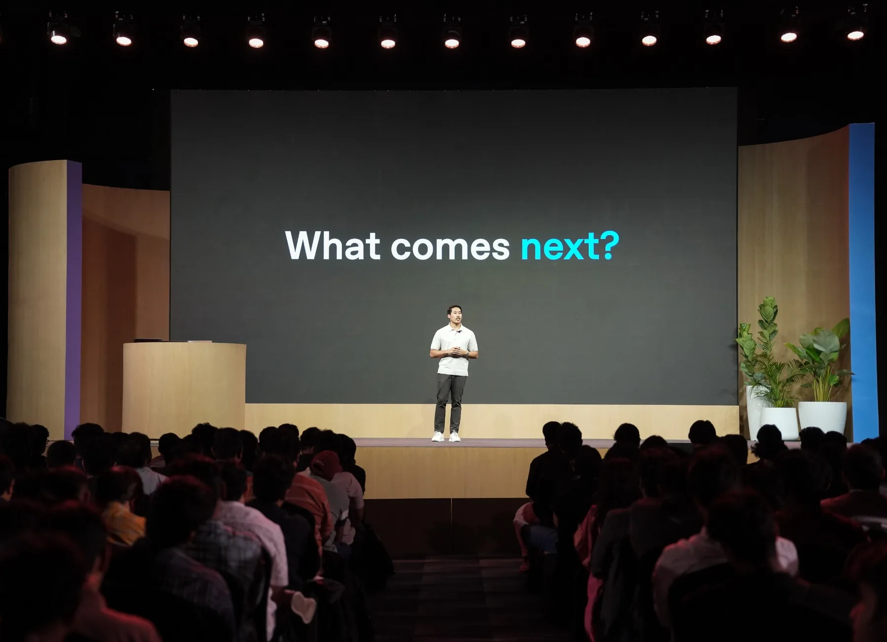 Derrick Choi presenting to an auditorium audience beneath a slide reading What comes next?